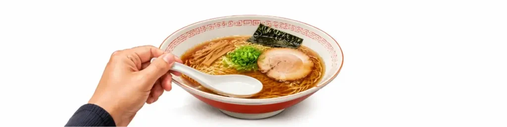白背景の横長画像。醤油ラーメンの入った丼が中央に置かれ、左手で白いレンゲの持ち手を支えている。レンゲの中は空で、レンゲの底がラーメンのスープ表面に触れる程度の低い位置に保たれている。丼の中には麺、チャーシュー、メンマ、海苔、刻みネギが見えており、レンゲを高く持ち上げず、スープ面近くで受け皿として構える状態を示している。